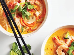 Orange broth filled with rice noodles, shredded carrots, sliced mushrooms, and tail-on cooked shrimp garnished with fresh cilantro